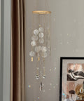 A hanging wind chime ornament with circular glass discs, gold-toned metal rods, and clear teardrop prisms, suspended in a room next to a brown curtain and a framed print.