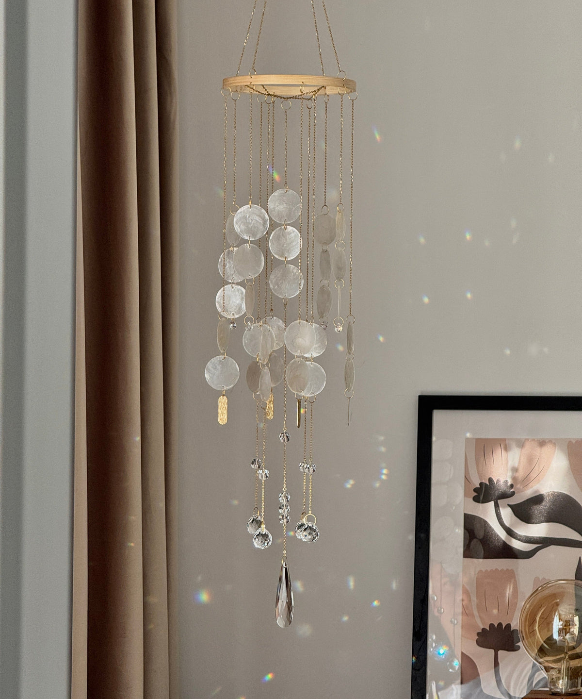 A hanging wind chime ornament with circular glass discs, gold-toned metal rods, and clear teardrop prisms, suspended in a room next to a brown curtain and a framed print.