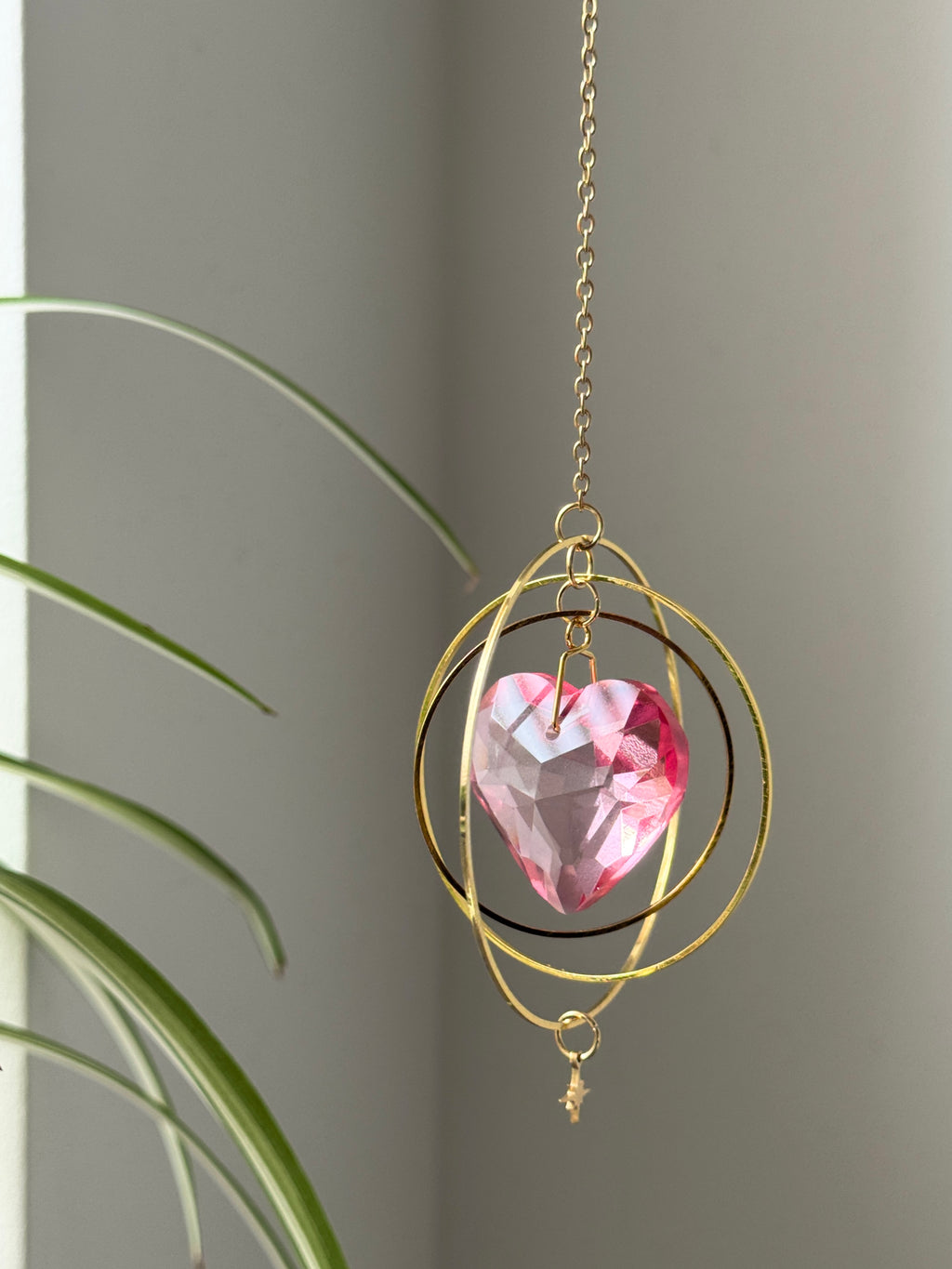 Sunkissed Heartbeat crystal suncatcher featuring a pink-gold faceted heart prism. Handmade celestial window decor with gold rings casting rainbow light flares (Sonnenfänger).