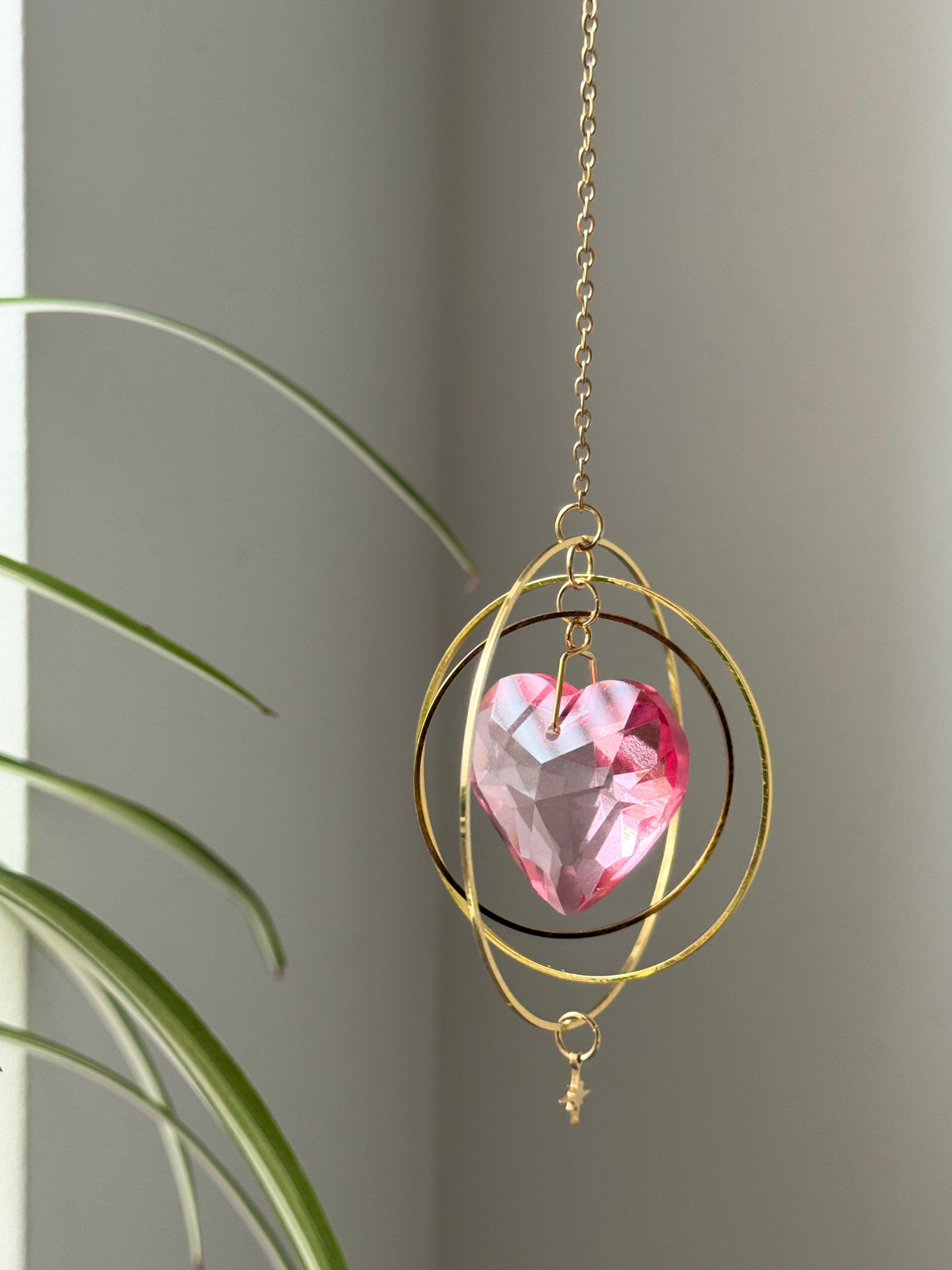 Sunkissed Heartbeat crystal suncatcher featuring a pink-gold faceted heart prism. Handmade celestial window decor with gold rings casting rainbow light flares (Sonnenfänger).