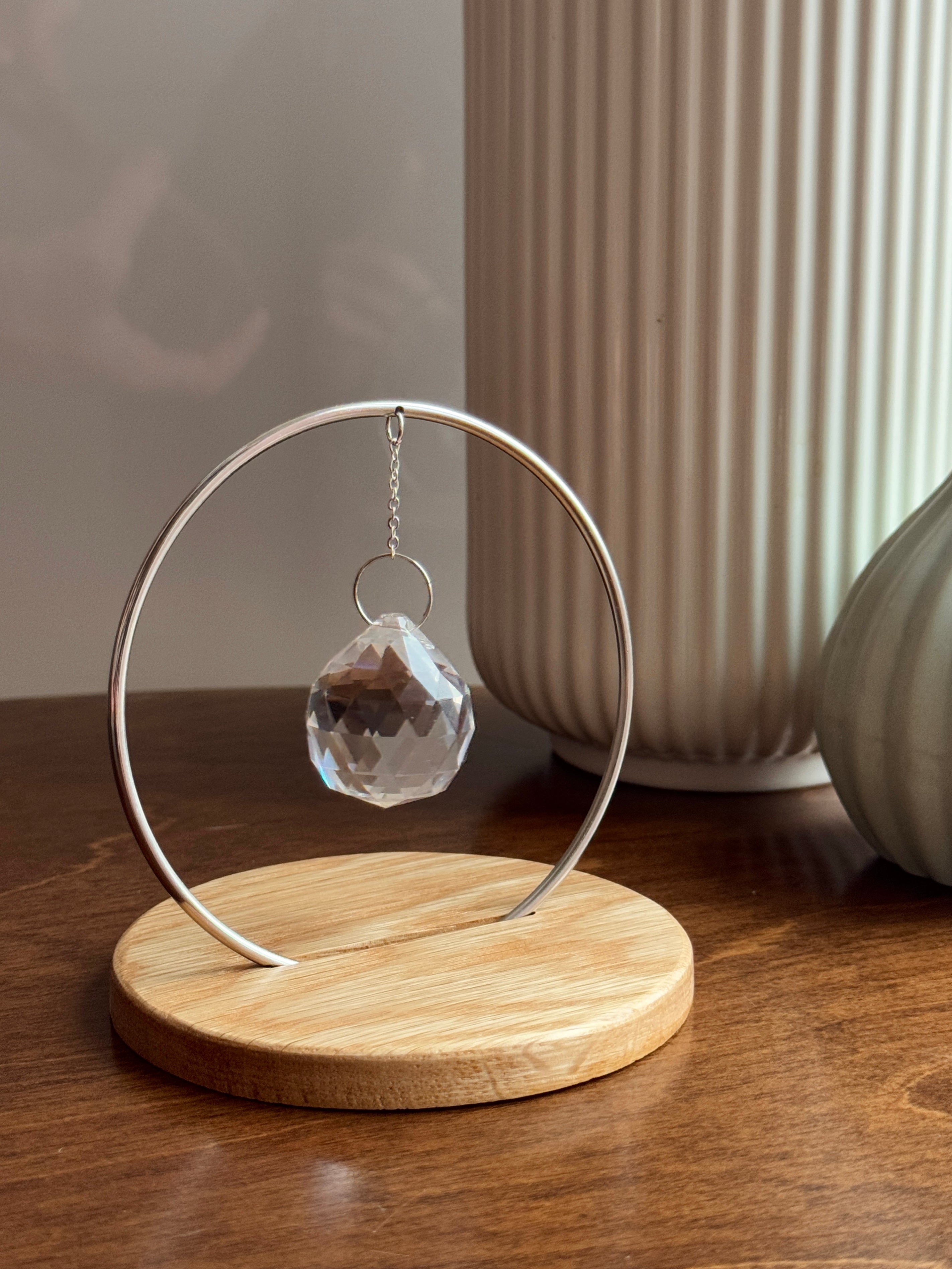 Minimalist "The Circle" tabletop suncatcher featuring a faceted crystal prism hanging inside a stainless steel ring on a round American oak base, casting vibrant rainbows on a white wall.