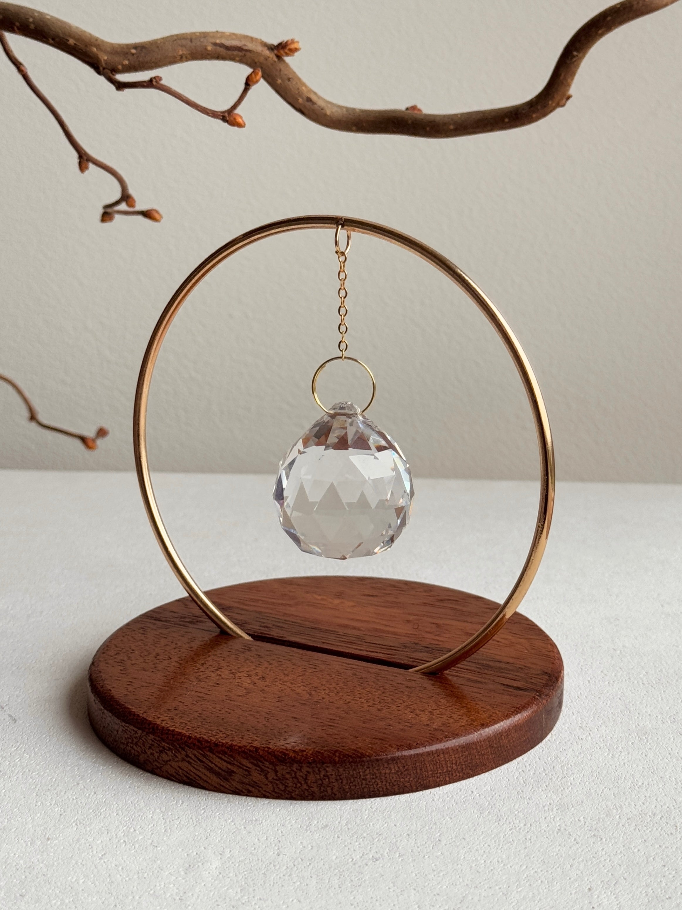 The Circle - A minimalist Sonnenfänger (suncatcher) featuring a high-quality faceted Kristall-Prisma suspended by a delicate gold stainless steel chain within a matching gold ring. The modern tabletop design rests on a solid, dark redwood base (Echtholz-Sockel) and is shown creating vibrant rainbow light reflections (Regenbogen-Effekt) across the background. This elegant Fensterdeko is perfect as a mindful Geschenkidee for home or office.