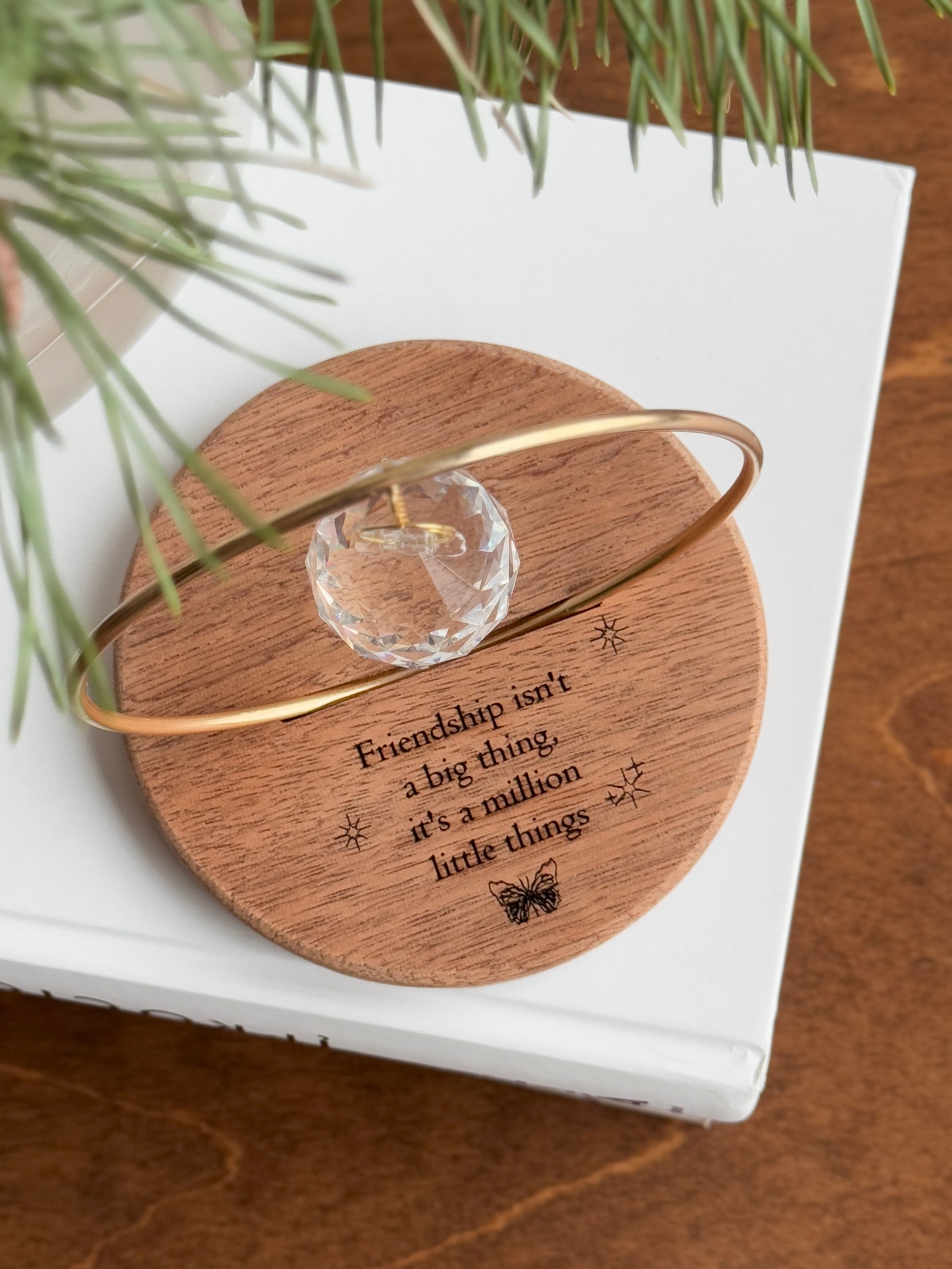 The Circle - A minimalist Sonnenfänger (suncatcher) featuring a high-quality faceted Kristall-Prisma suspended by a delicate gold stainless steel chain within a matching gold ring. The modern tabletop design rests on a solid, dark redwood base (Echtholz-Sockel) and is shown creating vibrant rainbow light reflections (Regenbogen-Effekt) across the background. This elegant Fensterdeko is perfect as a mindful Geschenkidee for home or office.