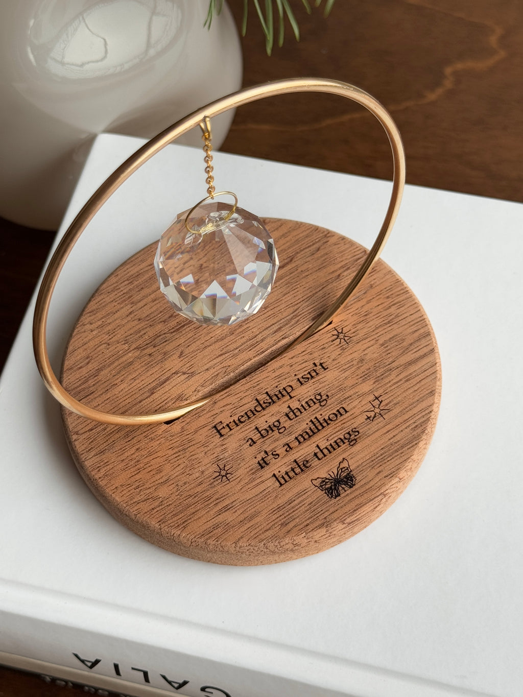 The Circle - A minimalist Sonnenfänger (suncatcher) featuring a high-quality faceted Kristall-Prisma suspended by a delicate gold stainless steel chain within a matching gold ring. The modern tabletop design rests on a solid, dark redwood base (Echtholz-Sockel) and is shown creating vibrant rainbow light reflections (Regenbogen-Effekt) across the background. This elegant Fensterdeko is perfect as a mindful Geschenkidee for home or office.