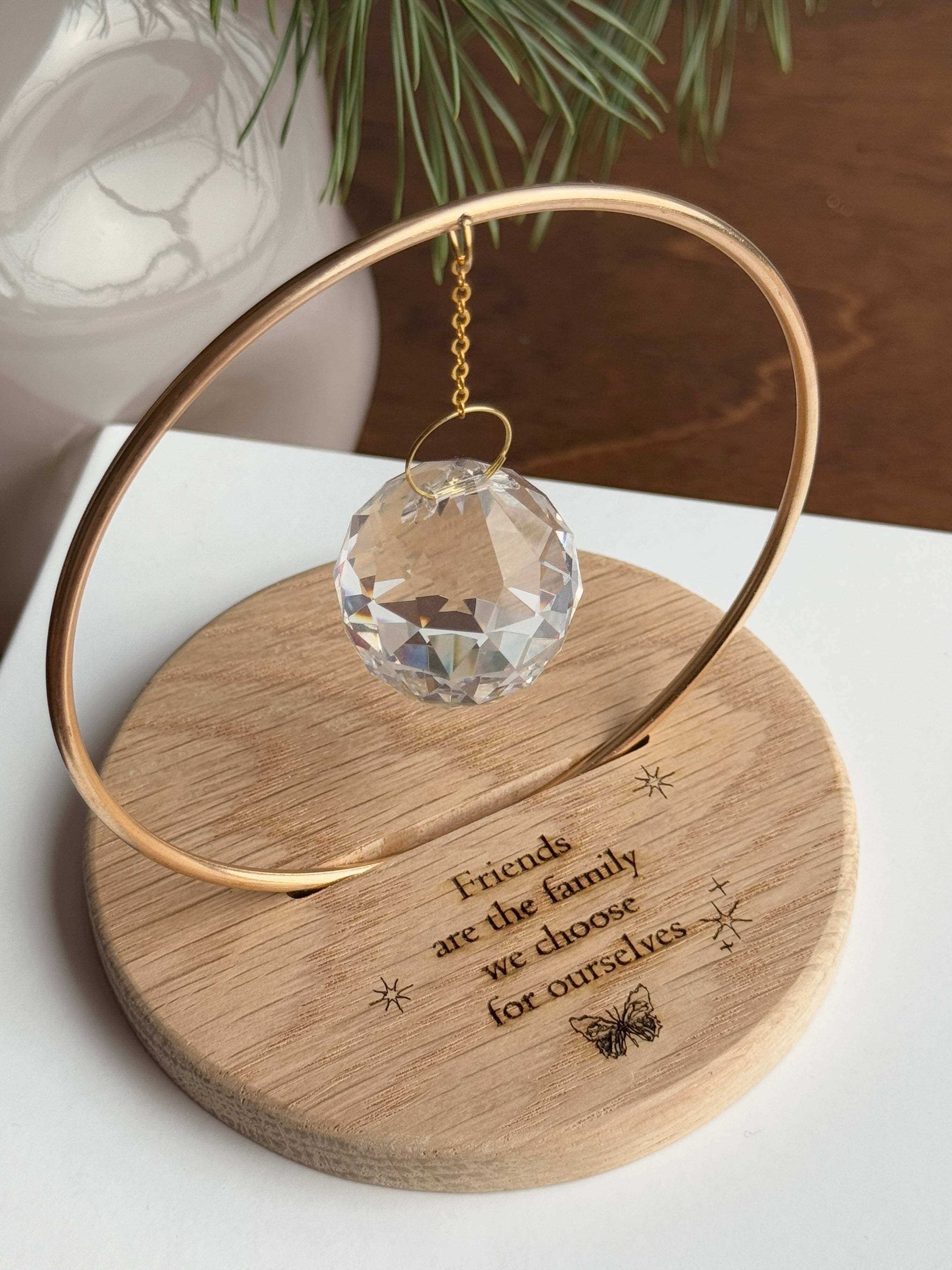 The Circle - A minimalist Sonnenfänger (suncatcher) featuring a high-quality faceted Kristall-Prisma suspended by a delicate gold stainless steel chain within a matching gold ring. The modern tabletop design rests on a solid, dark redwood base (Echtholz-Sockel) and is shown creating vibrant rainbow light reflections (Regenbogen-Effekt) across the background. This elegant Fensterdeko is perfect as a mindful Geschenkidee for home or office.