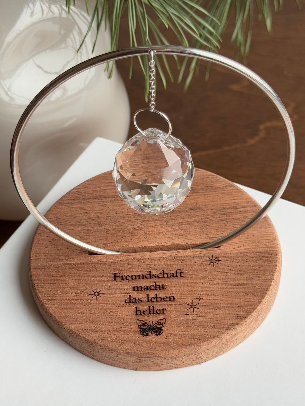 The Circle - A minimalist Sonnenfänger (suncatcher) featuring a high-quality faceted Kristall-Prisma suspended by a delicate gold stainless steel chain within a matching gold ring. The modern tabletop design rests on a solid, dark redwood base (Echtholz-Sockel) and is shown creating vibrant rainbow light reflections (Regenbogen-Effekt) across the background. This elegant Fensterdeko is perfect as a mindful Geschenkidee for home or office.