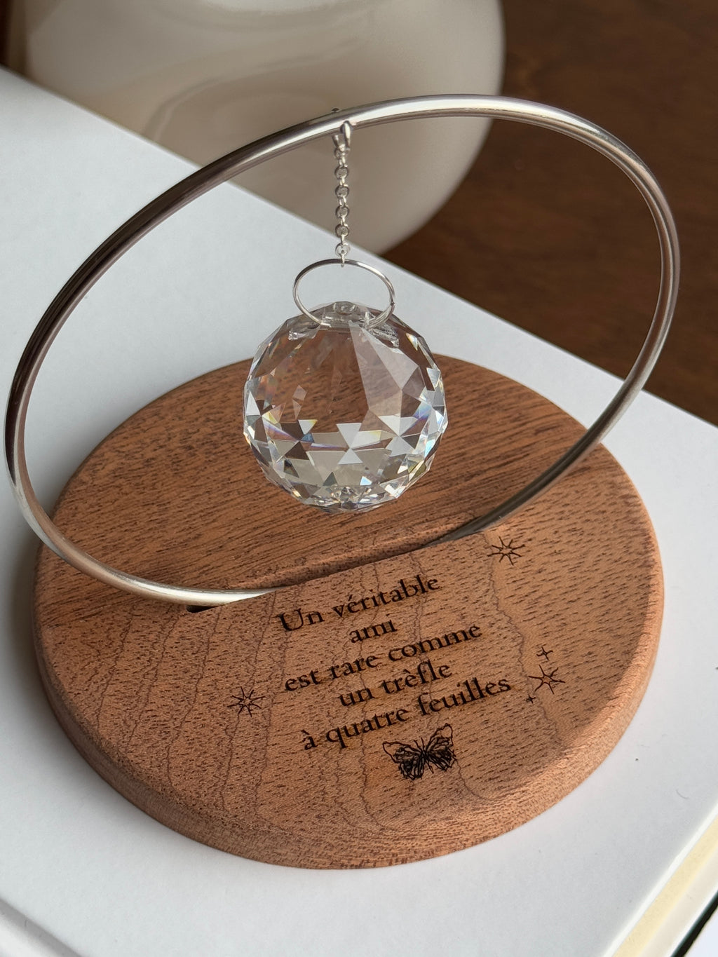 The Circle - A minimalist Sonnenfänger (suncatcher) featuring a high-quality faceted Kristall-Prisma suspended by a delicate gold stainless steel chain within a matching gold ring. The modern tabletop design rests on a solid, dark redwood base (Echtholz-Sockel) and is shown creating vibrant rainbow light reflections (Regenbogen-Effekt) across the background. This elegant Fensterdeko is perfect as a mindful Geschenkidee for home or office.