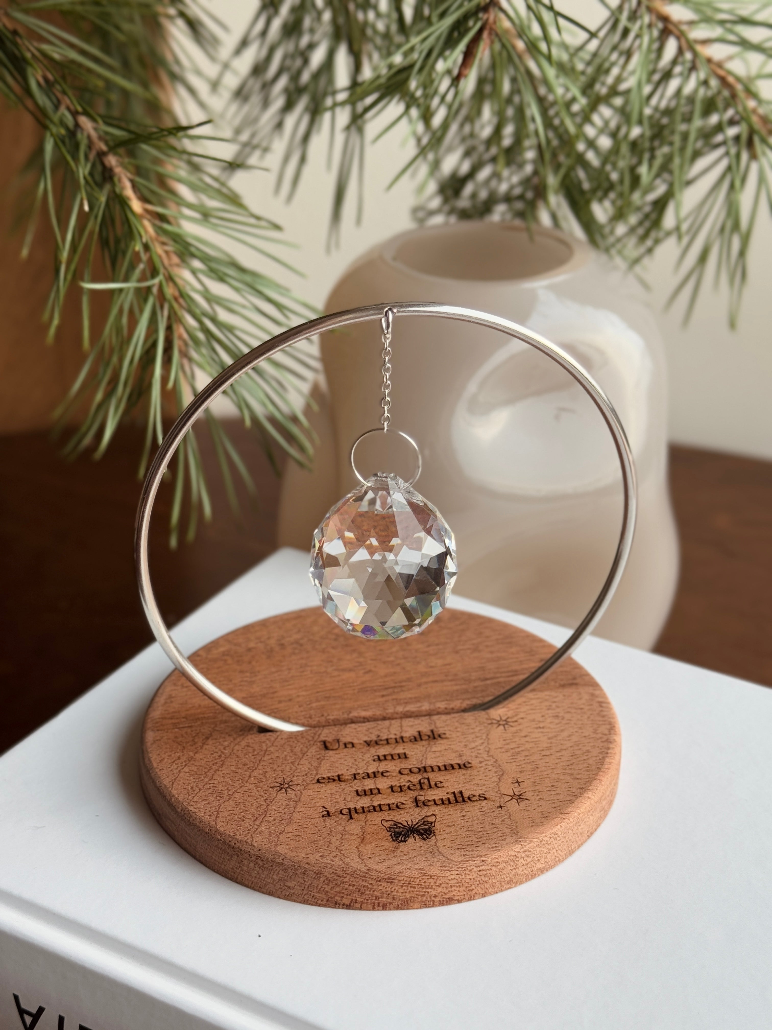 The Circle - A minimalist Sonnenfänger (suncatcher) featuring a high-quality faceted Kristall-Prisma suspended by a delicate gold stainless steel chain within a matching gold ring. The modern tabletop design rests on a solid, dark redwood base (Echtholz-Sockel) and is shown creating vibrant rainbow light reflections (Regenbogen-Effekt) across the background. This elegant Fensterdeko is perfect as a mindful Geschenkidee for home or office.