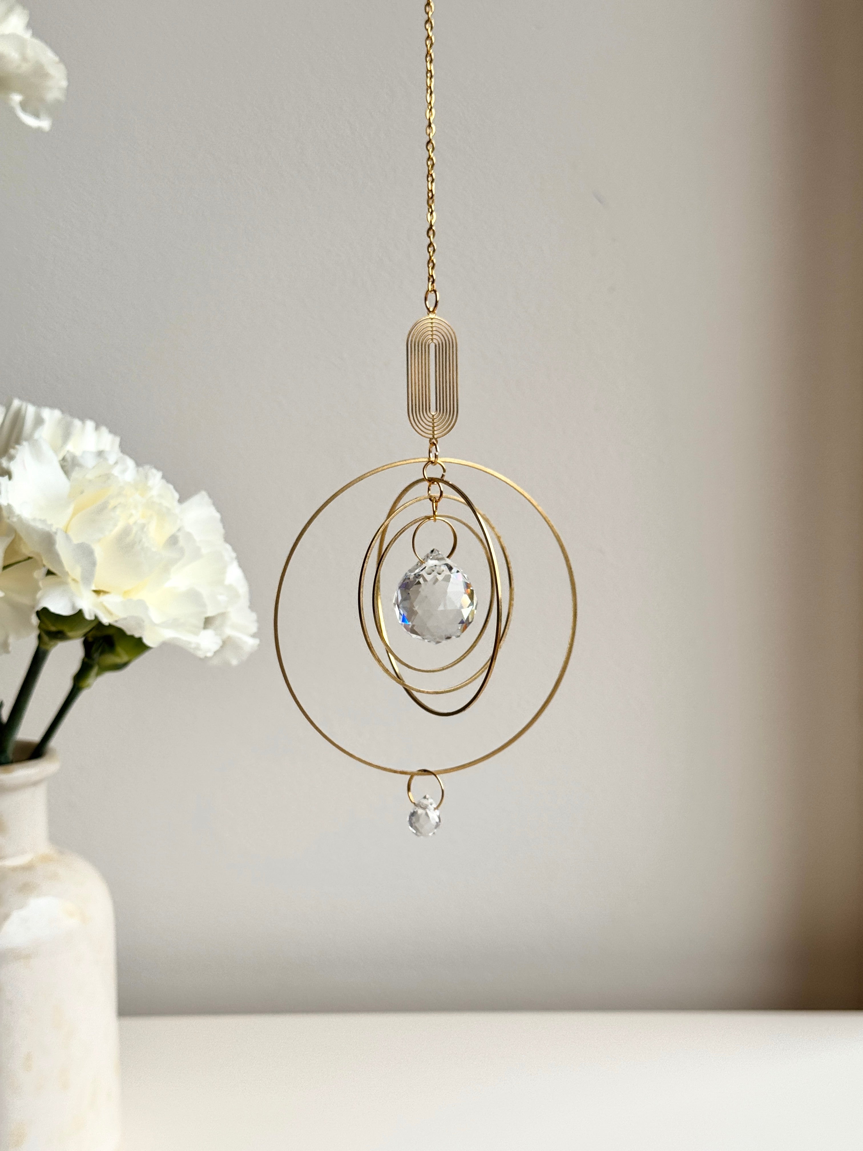 Close-up of a handmade golden boho suncatcher (Sonnenfänger) with multiple concentric rings and a faceted crystal prism (Kristall), shown next to delicate white flowers.