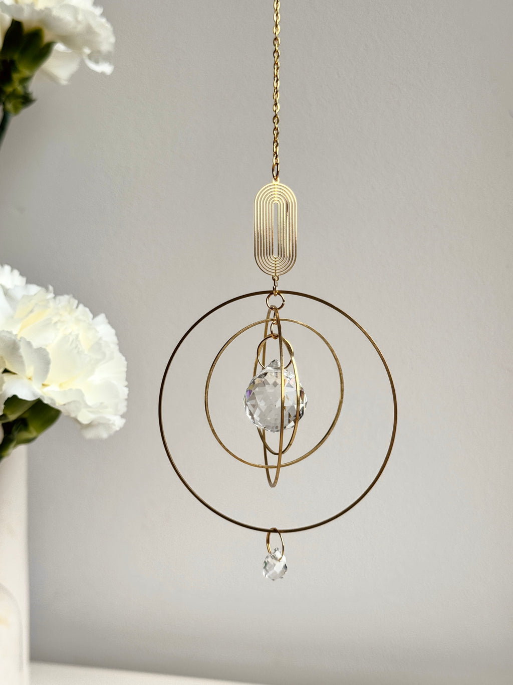 Close-up of a handmade golden boho suncatcher (Sonnenfänger) with multiple concentric rings and a faceted crystal prism (Kristall), shown next to delicate white flowers.