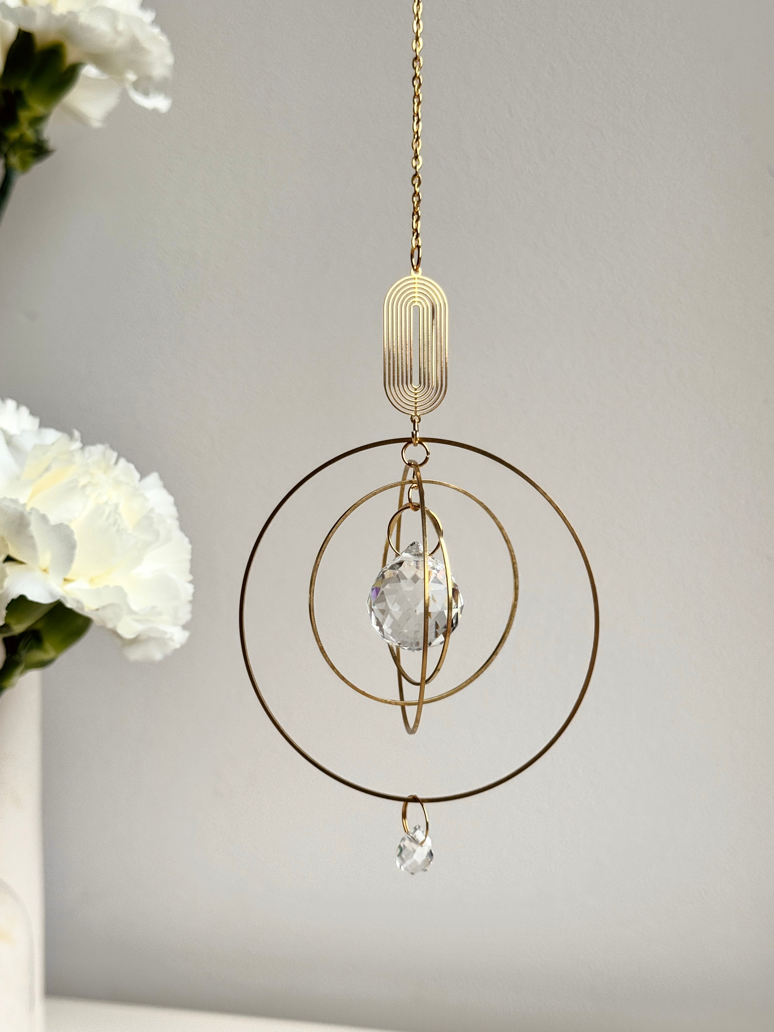 Close-up of a handmade golden boho suncatcher (Sonnenfänger) with multiple concentric rings and a faceted crystal prism (Kristall), shown next to delicate white flowers.