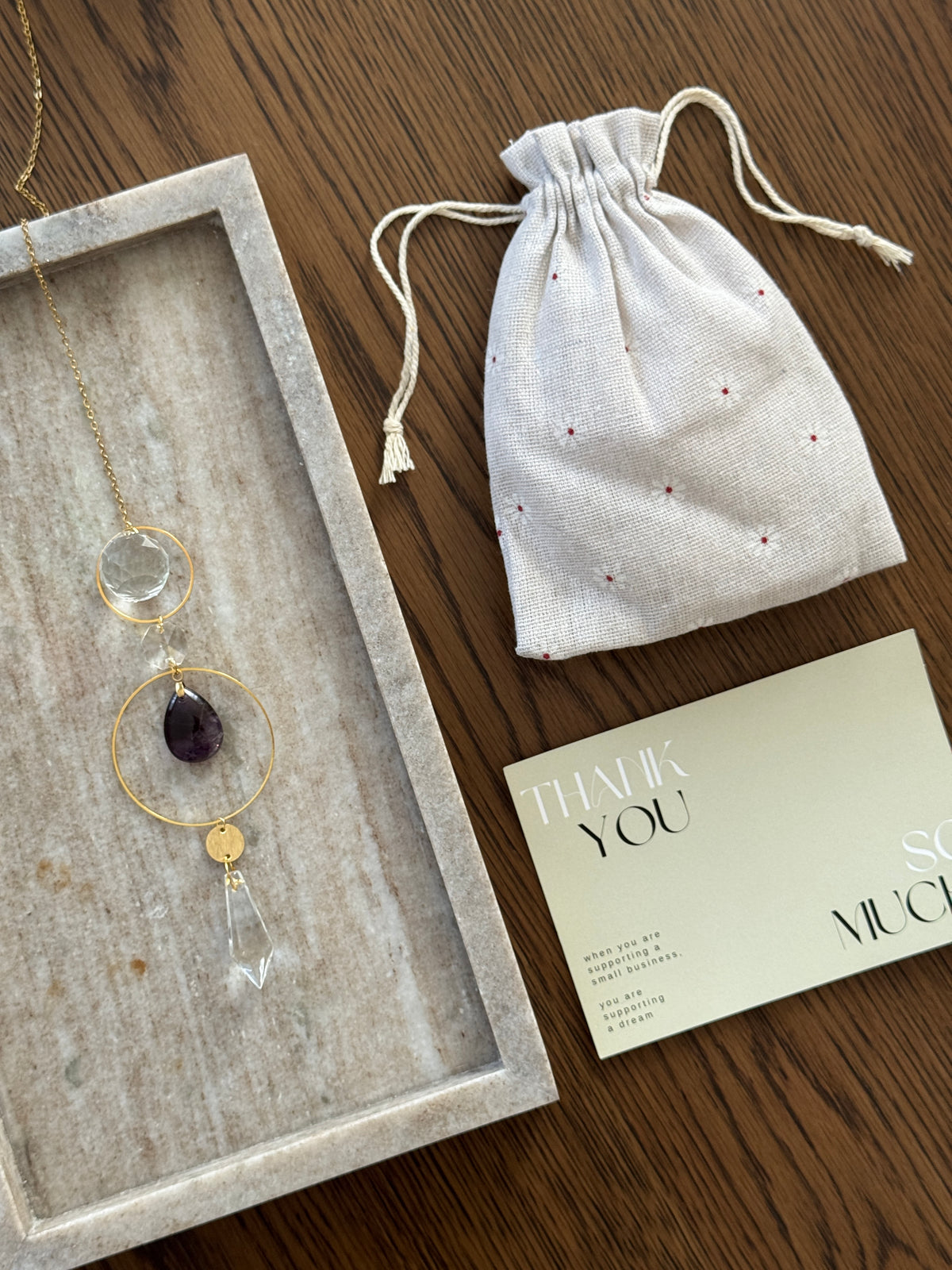 A gold-toned necklace with a clear teardrop crystal and purple amethyst pendant is displayed in a gray tray next to a white drawstring pouch and a thank you card on a wooden table.