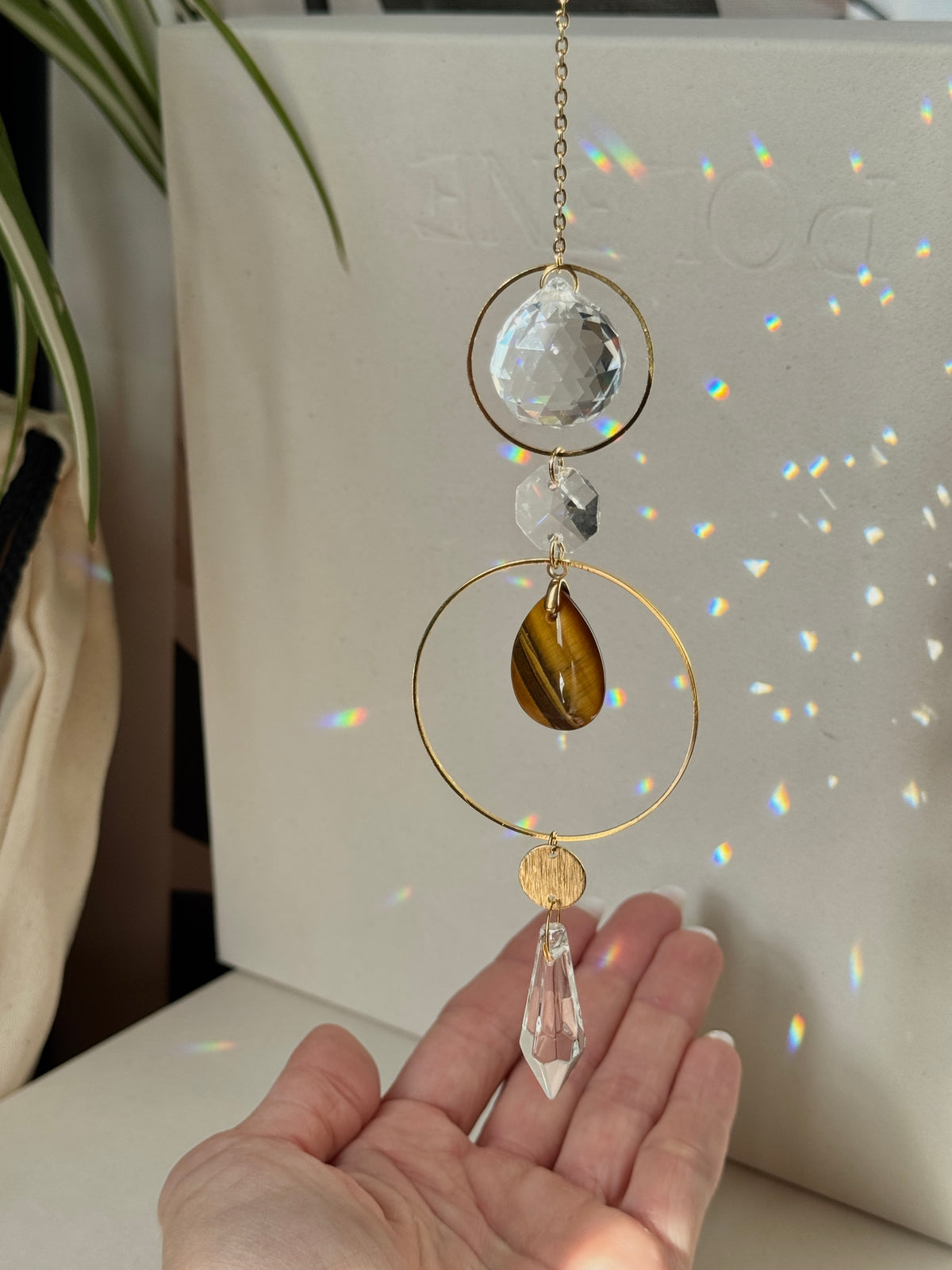 A gold and crystal suncatcher ornament with a faceted clear top sphere, a small clear bead, a brown amber teardrop, a clear crystal drop, and a clear circular ring, held in front of a white laptop by a hand.