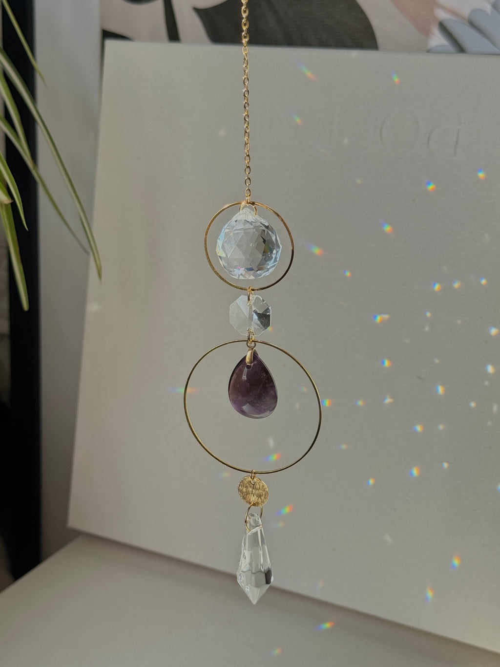 A gold-toned chain suncatcher ornament featuring a faceted clear crystal, a purple teardrop amethyst, and a clear crystal point, displayed against a light gray backdrop with rainbow light reflections.
