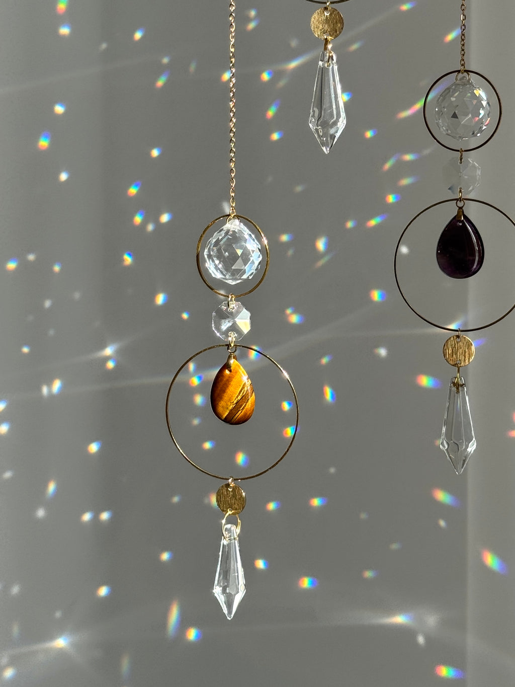 Gold-toned circular suncatcher ornaments with clear crystal prisms and a tiger's eye stone, hanging against a wall with rainbow light projections.