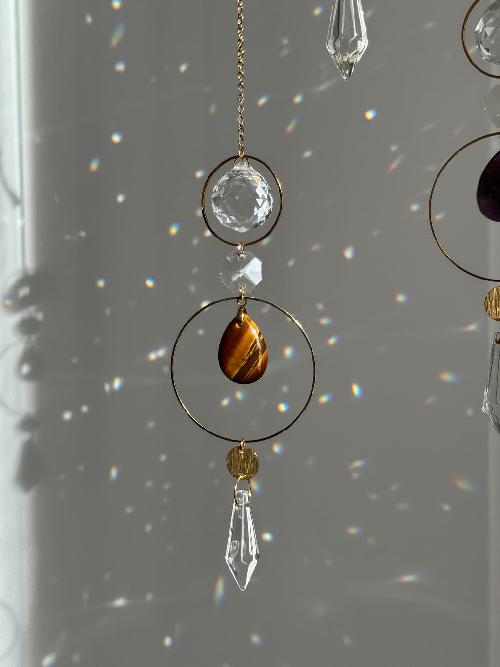 A close-up of a hanging decorative ornament with gold-tone rings, clear faceted crystal prisms, and a teardrop-shaped brown tiger's eye stone, shown against a gray wall with light rainbows reflecting on it.