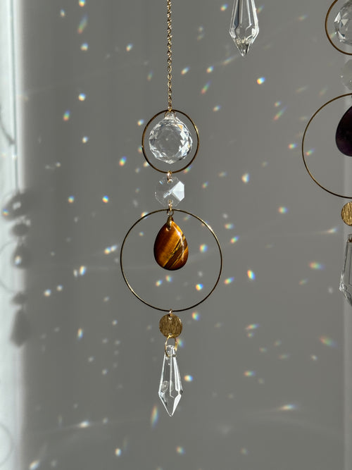A close-up of a hanging decorative ornament with gold-tone rings, clear faceted crystal prisms, and a teardrop-shaped brown tiger's eye stone, shown against a gray wall with light rainbows reflecting on it.