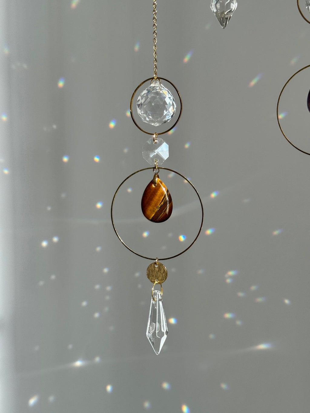 A hanging decorative ornament with a gold-tone chain, clear faceted crystal beads, a small clear crystal cube, a gold ring, a brown tiger's eye teardrop, a small gold disc, and a clear crystal prism dangling below, set against a soft gray background with light rainbows.