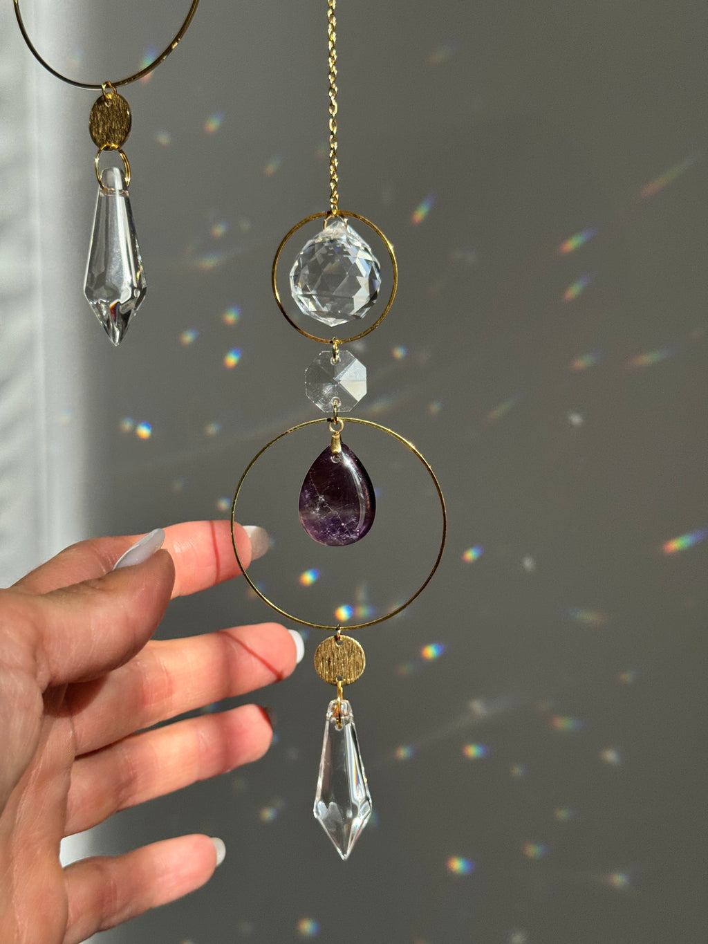 A gold-toned crystal suncatcher ornament with a purple amethyst teardrop, clear faceted crystal drops, and a small clear crystal bead, held by a hand against a gray wall with sunlight casting rainbow reflections.