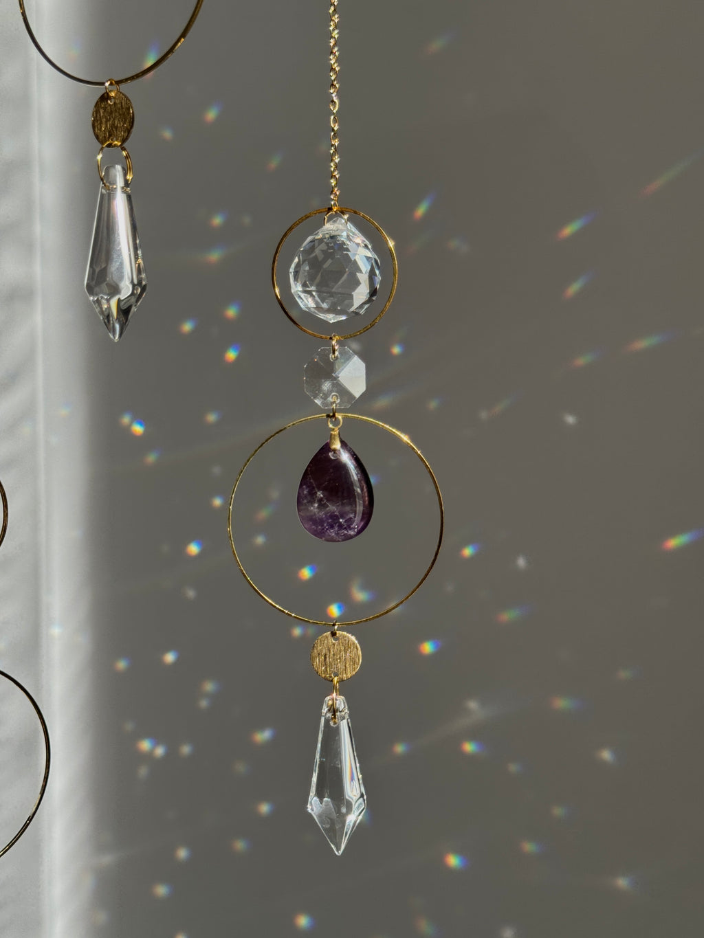 A decorative hanging ornament with gold-tone rings and chains, featuring a faceted clear crystal teardrop, a clear oval crystal, a small clear bead, and a purple amethyst teardrop, shown against a gray wall with rainbow light reflections.