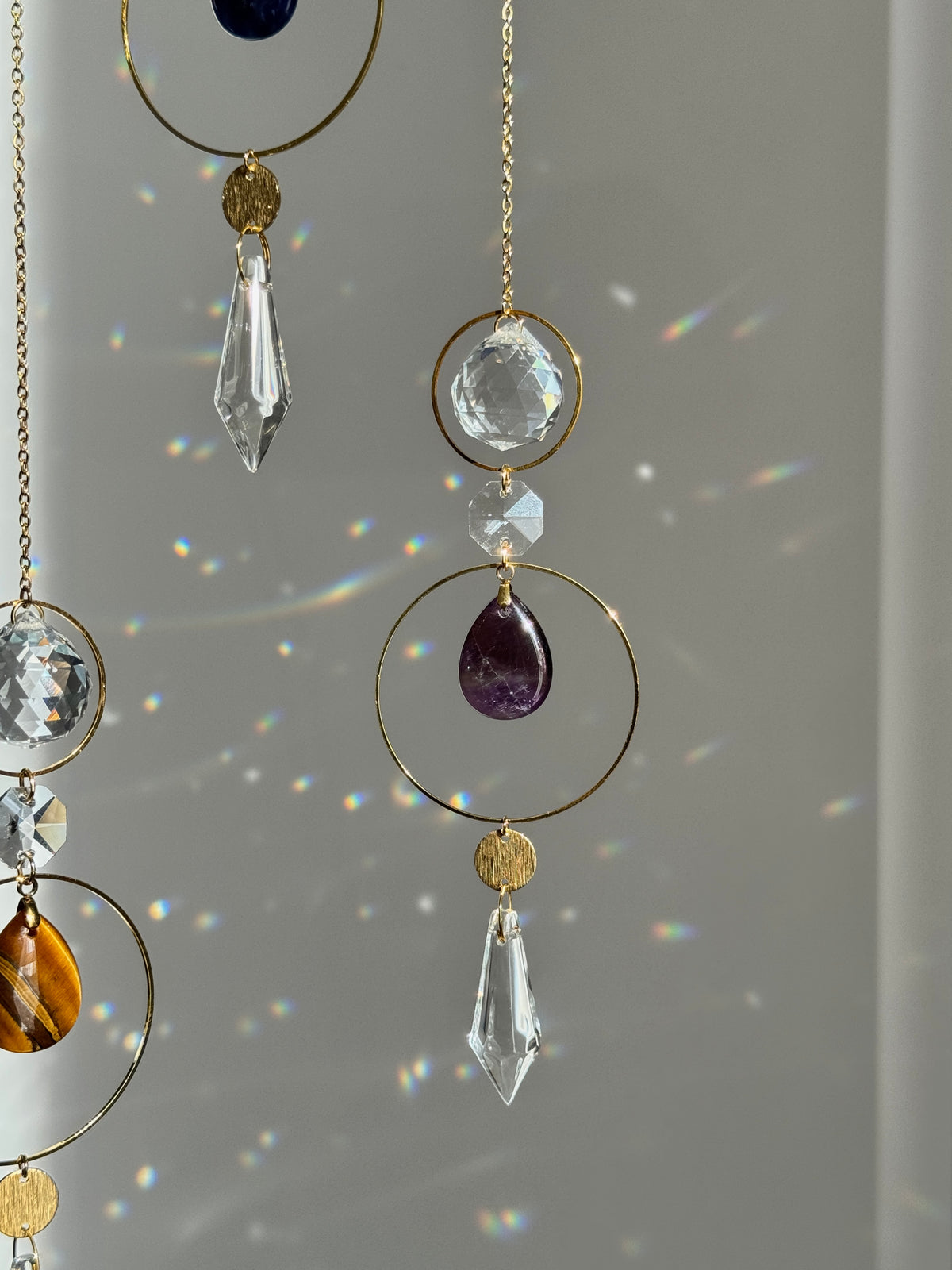 Gold-tone suncatcher mobile with clear crystal prisms, a purple teardrop gemstone, and amber crystal accents, shown hanging against a window with sunlight and rainbow reflections.