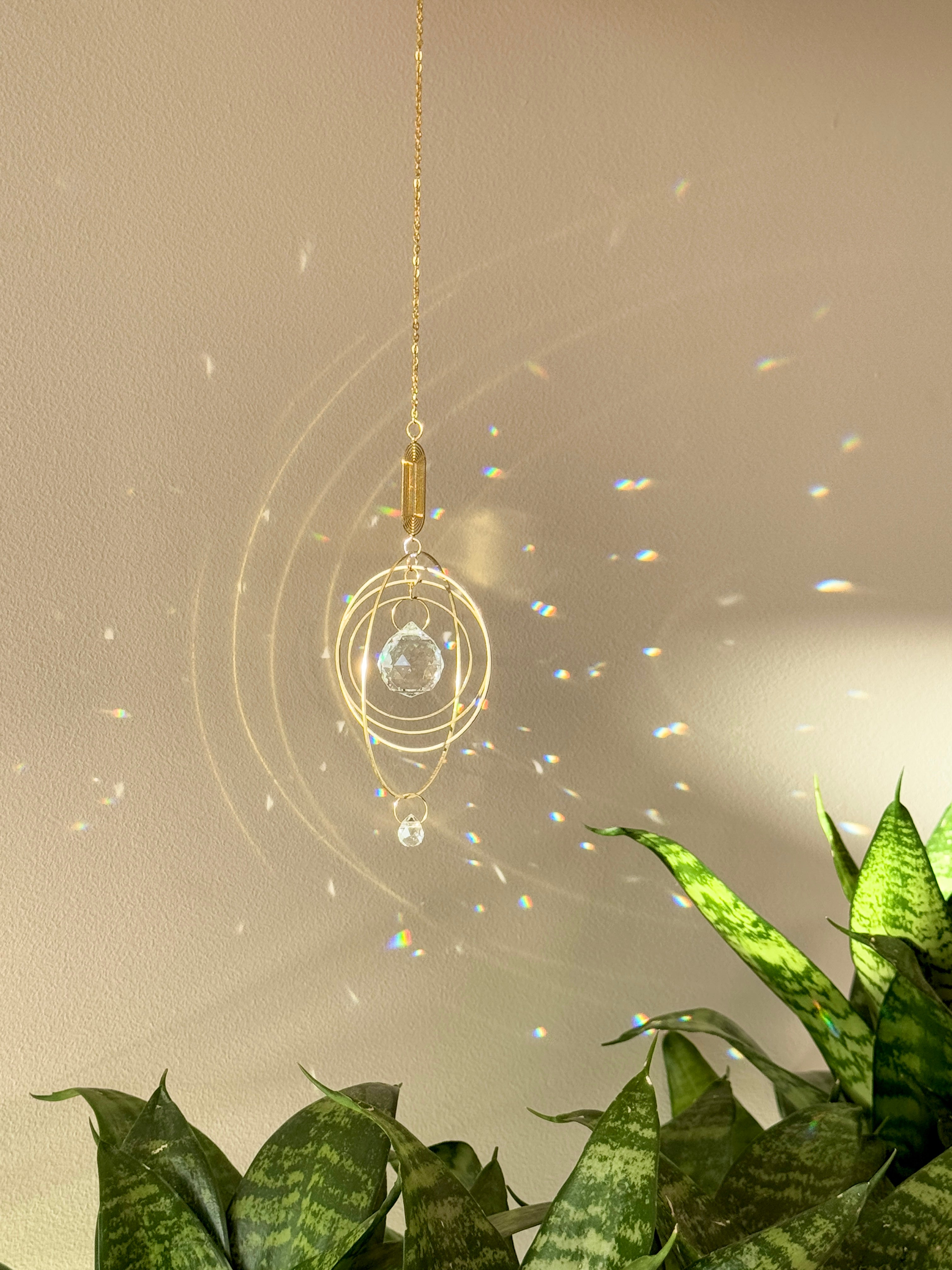 Celestial boho window suncatcher (Sonnenfänger Fenster) with gold concentric rings and a crystal prism, scattering magical rainbows (Regenbogen) across a sunlit wall next to a Monstera leaf.