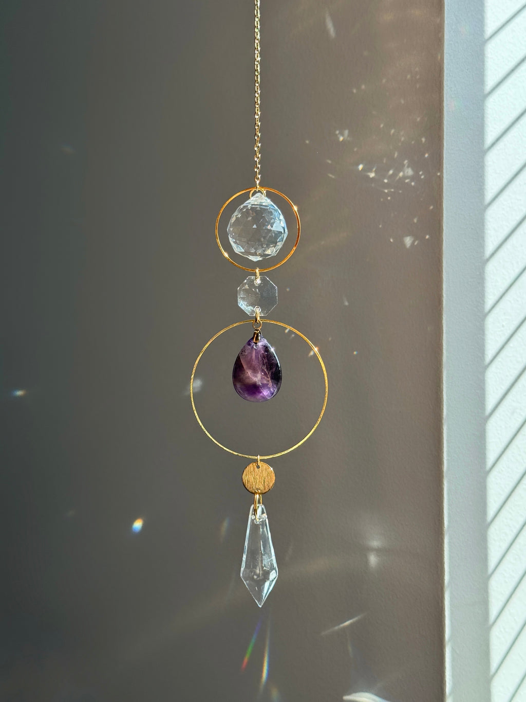 A gold-toned hanging suncatcher with a clear faceted crystal drop, a purple teardrop gemstone, and clear round beads, photographed in front of a window with sunlight and lens flares.