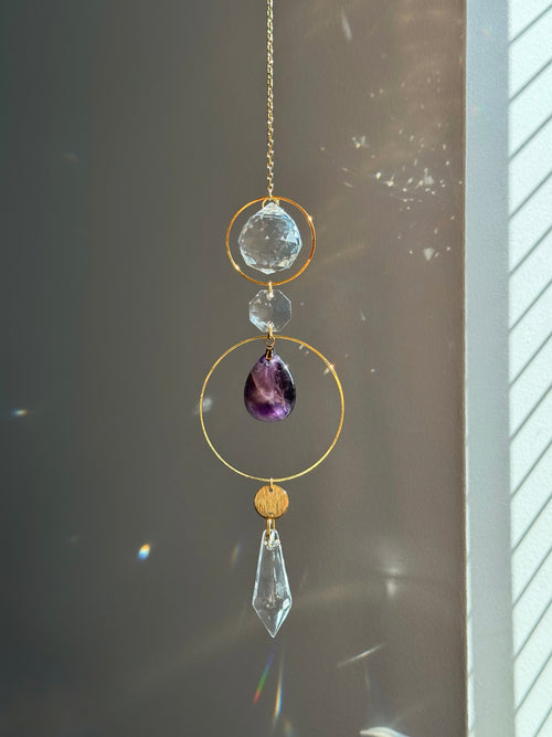 A gold-toned hanging suncatcher with a clear faceted crystal drop, a purple teardrop gemstone, and clear round beads, photographed in front of a window with sunlight and lens flares.