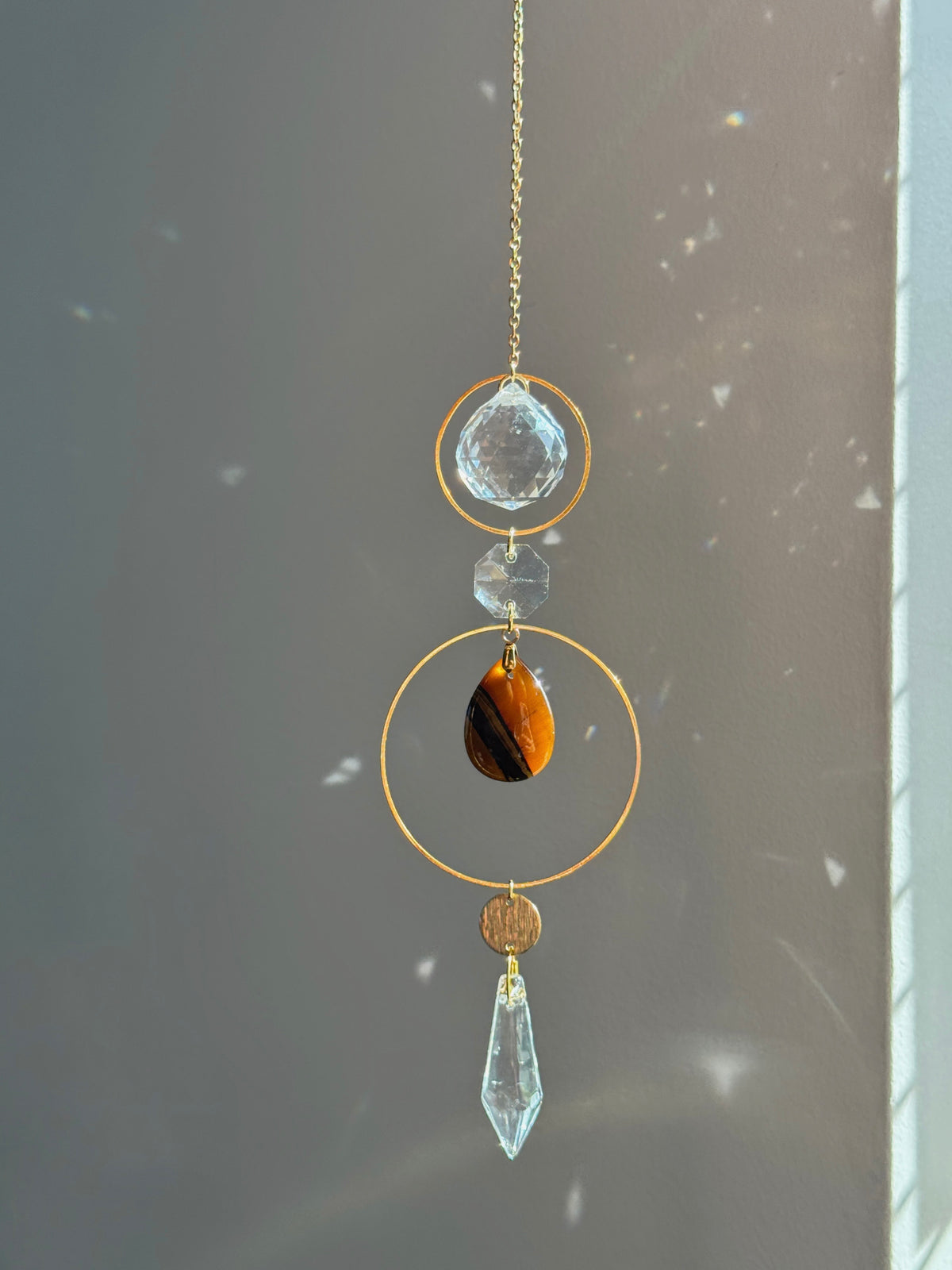 A gold-toned hanging suncatcher ornament with a clear faceted crystal, a small clear bead, a brown tiger's eye stone, and a clear teardrop crystal, shown against a gray wall with sunlight and lens flare effects.