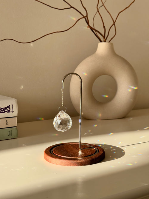 A close-up photograph of a clear, multi-faceted crystal suncatcher prism hanging from an ornate rose gold metal hook on a dark wooden base. The suncatcher casts multiple rainbow-colored refractions of light across the white tabletop, a stack of white books on the left, and a textured off-white ceramic vase in the background. Dried branches are visible emerging from the vase against a light gray wall.