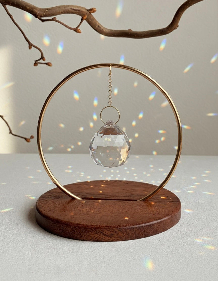 The Circle - A minimalist Sonnenfänger (suncatcher) featuring a high-quality faceted Kristall-Prisma suspended by a delicate gold stainless steel chain within a matching gold ring. The modern tabletop design rests on a solid, dark redwood base (Echtholz-Sockel) and is shown creating vibrant rainbow light reflections (Regenbogen-Effekt) across the background. This elegant Fensterdeko is perfect as a mindful Geschenkidee for home or office.