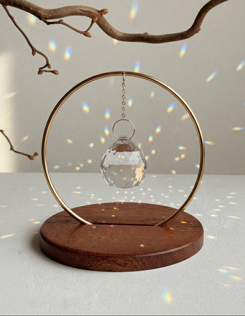 The Circle - A minimalist Sonnenfänger (suncatcher) featuring a high-quality faceted Kristall-Prisma suspended by a delicate gold stainless steel chain within a matching gold ring. The modern tabletop design rests on a solid, dark redwood base (Echtholz-Sockel) and is shown creating vibrant rainbow light reflections (Regenbogen-Effekt) across the background. This elegant Fensterdeko is perfect as a mindful Geschenkidee for home or office.