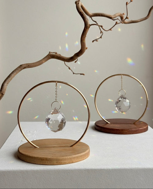 The Circle - A minimalist Sonnenfänger (suncatcher) featuring a high-quality faceted Kristall-Prisma suspended by a delicate gold stainless steel chain within a matching gold ring. The modern tabletop design rests on a solid, dark redwood base (Echtholz-Sockel) and is shown creating vibrant rainbow light reflections (Regenbogen-Effekt) across the background. This elegant Fensterdeko is perfect as a mindful Geschenkidee for home or office.