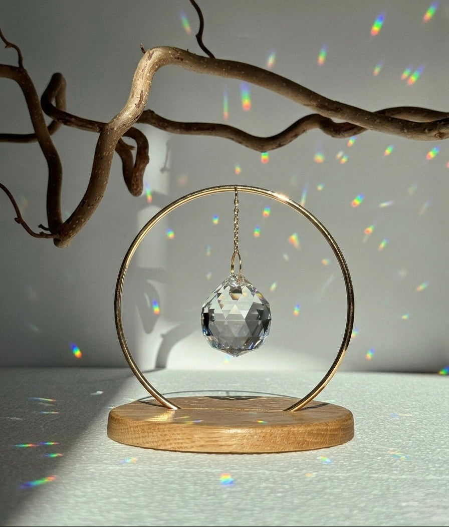A clear, faceted crystal prism sphere hangs by a delicate gold chain within a circular brass frame, titled "The Circle." The suncatcher is mounted on a smooth, light oak wooden base. Sunlight passes through the crystal, scattering vibrant rainbow light leaks and spectrum dots across a white background and onto gnarled, aesthetic tree branches. The scene is bright, minimalist, and serene.