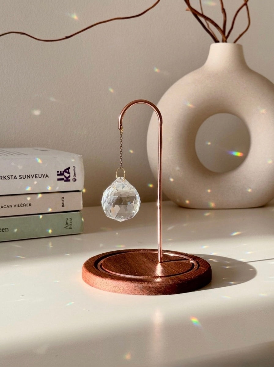 A close-up photograph of a clear, multi-faceted crystal suncatcher prism hanging from an ornate rose gold metal hook on a dark wooden base. The suncatcher casts multiple rainbow-colored refractions of light across the white tabletop, a stack of white books on the left, and a textured off-white ceramic vase in the background. Dried branches are visible emerging from the vase against a light gray wall.