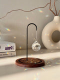 The Aura Hook tabletop suncatcher featuring a faceted crystal prism hanging from a black minimalist stand on a round redwood base, casting rainbow light reflections on a white surface.