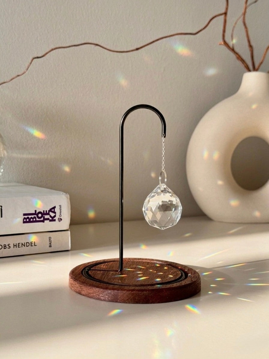 The Aura Hook tabletop suncatcher featuring a faceted crystal prism hanging from a black minimalist stand on a round redwood base, casting rainbow light reflections on a white surface.