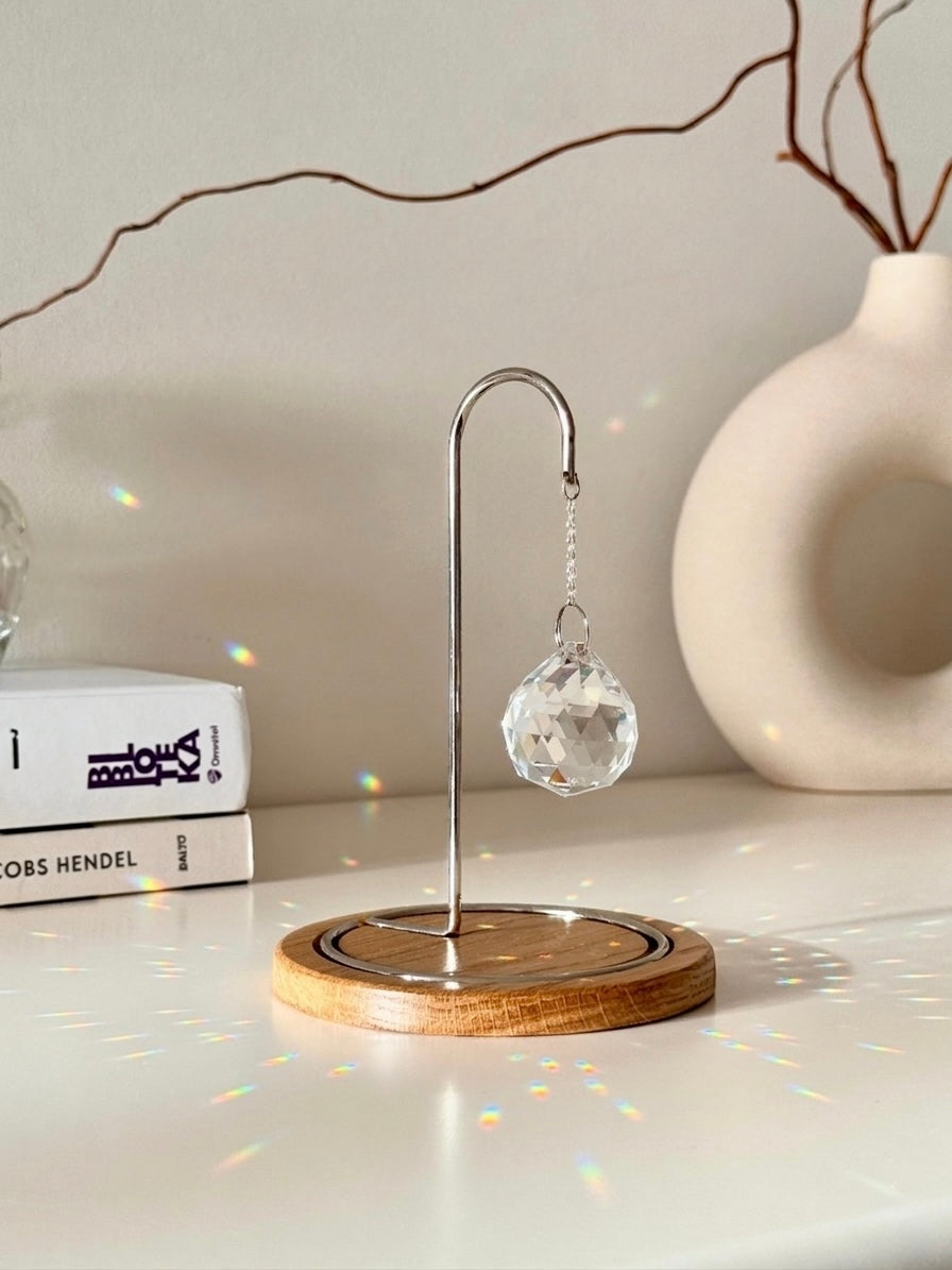 A close-up photograph of a clear, multi-faceted crystal suncatcher prism hanging from an ornate rose gold metal hook on a dark wooden base. The suncatcher casts multiple rainbow-colored refractions of light across the white tabletop, a stack of white books on the left, and a textured off-white ceramic vase in the background. Dried branches are visible emerging from the vase against a light gray wall.