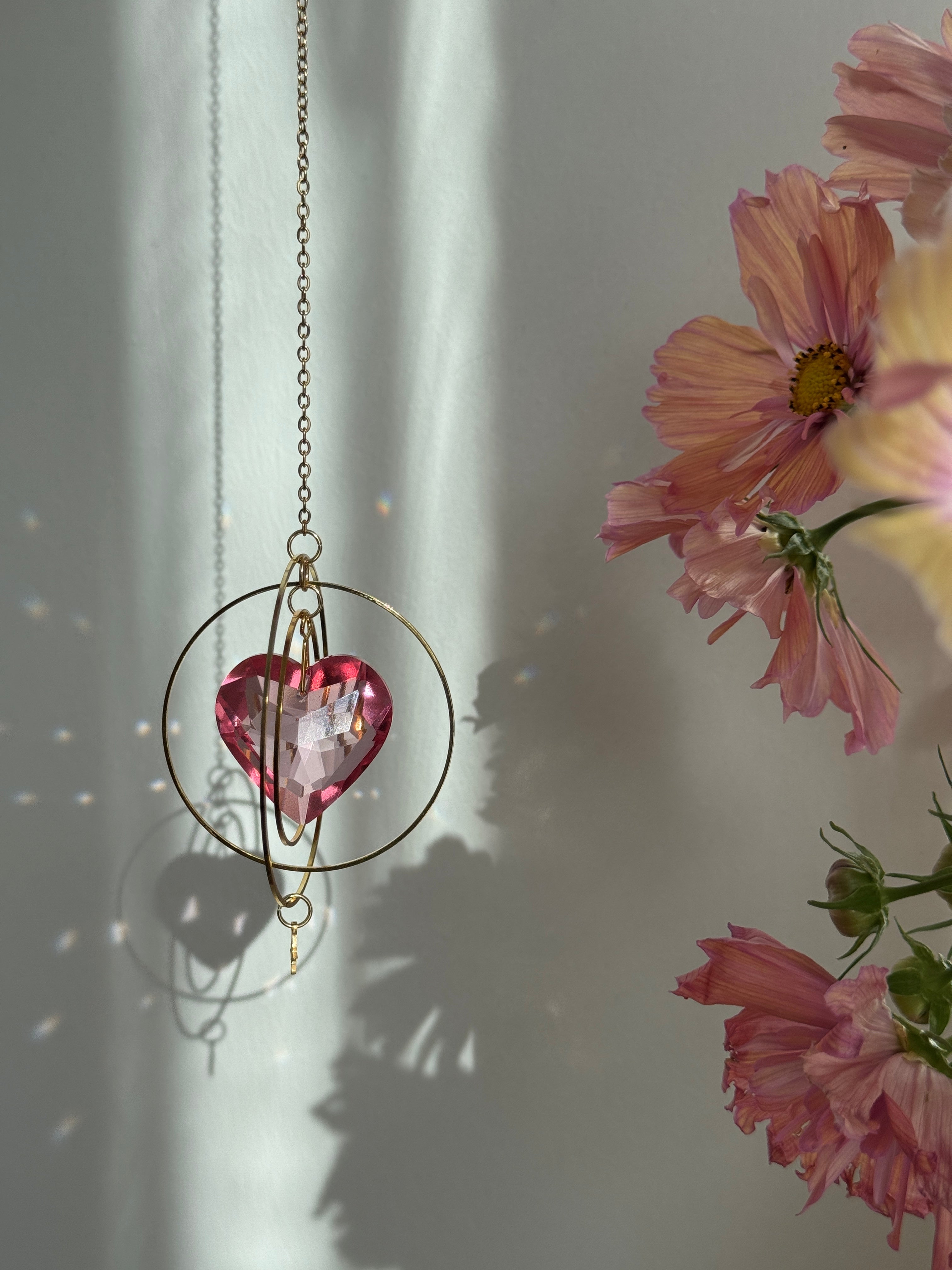 Sunkissed Heartbeat crystal suncatcher featuring a pink-gold faceted heart prism. Handmade celestial window decor with gold rings casting rainbow light flares (Sonnenfänger).