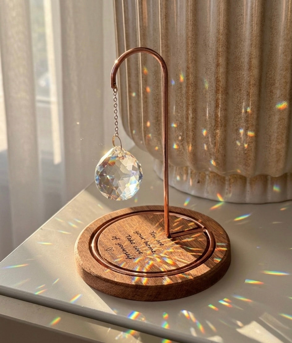 A faceted crystal suncatcher hangs from a rose gold metal hook on a circular wooden base. Sunlight streams through the crystal, scattering vibrant rainbow-colored light reflections across the base and the surrounding white surface. The wooden base is engraved with the message: "You are your home. Take care of yourself." followed by a small hand-drawn heart.