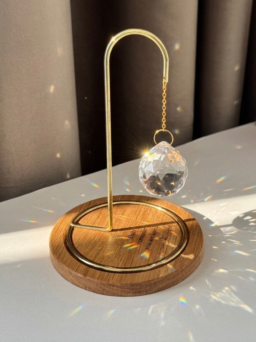 A faceted crystal prism hangs from a gold-toned metal hook over a light oak wooden base. Bright sunlight hits the crystal, creating a brilliant flare and casting delicate rainbow specks across the wood and white tabletop. The base is engraved with the words: "Daily reminder: you are loved." beside a minimalist hand-drawn heart.