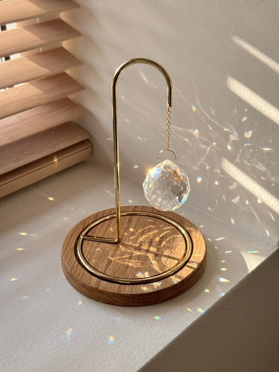 A faceted glass crystal sphere hangs from a gold wire stand on a circular wooden base, which is engraved with the words "Daily reminder: you are loved." The suncatcher is on a white surface next to a wooden window blind. Sunlight streams through the crystal, creating a radiant glow and casting intricate patterns of light and rainbow-colored reflections on the base and surrounding surfaces.