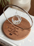 The Circle - A minimalist Sonnenfänger (suncatcher) featuring a high-quality faceted Kristall-Prisma suspended by a delicate gold stainless steel chain within a matching gold ring. The modern tabletop design rests on a solid, dark redwood base (Echtholz-Sockel) and is shown creating vibrant rainbow light reflections (Regenbogen-Effekt) across the background. This elegant Fensterdeko is perfect as a mindful Geschenkidee for home or office.