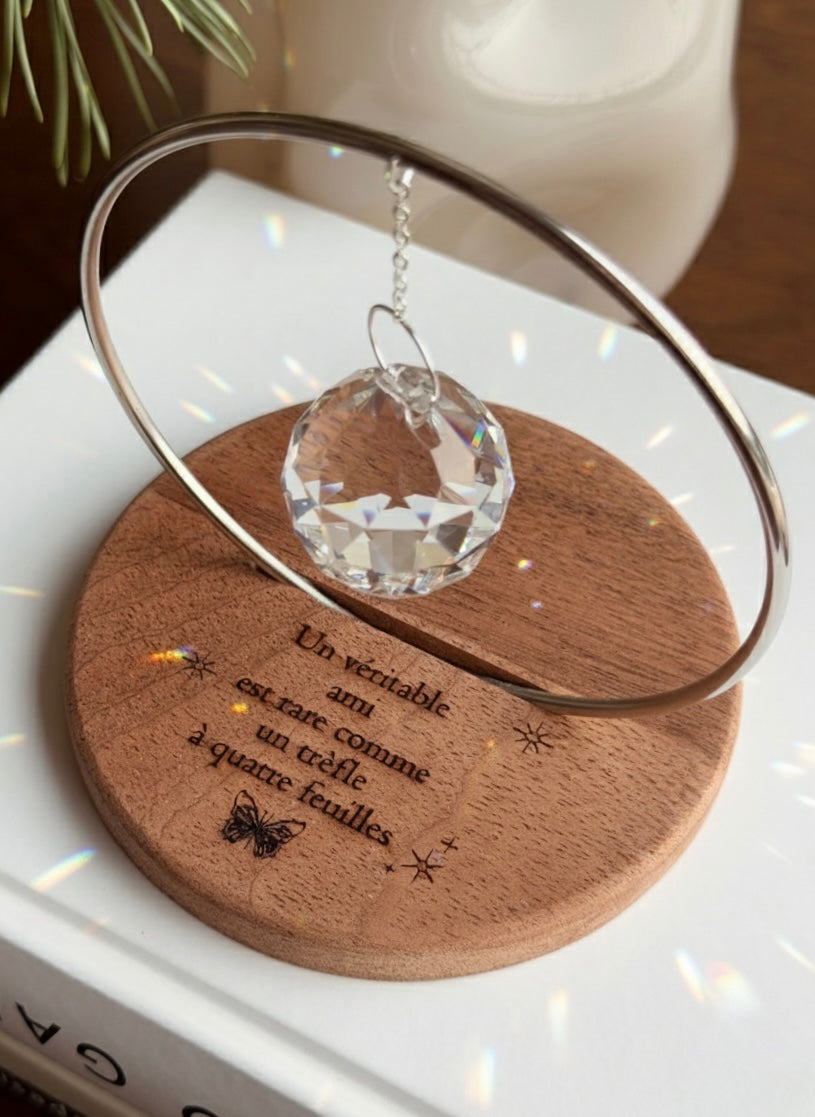 The Circle - A minimalist Sonnenfänger (suncatcher) featuring a high-quality faceted Kristall-Prisma suspended by a delicate gold stainless steel chain within a matching gold ring. The modern tabletop design rests on a solid, dark redwood base (Echtholz-Sockel) and is shown creating vibrant rainbow light reflections (Regenbogen-Effekt) across the background. This elegant Fensterdeko is perfect as a mindful Geschenkidee for home or office.