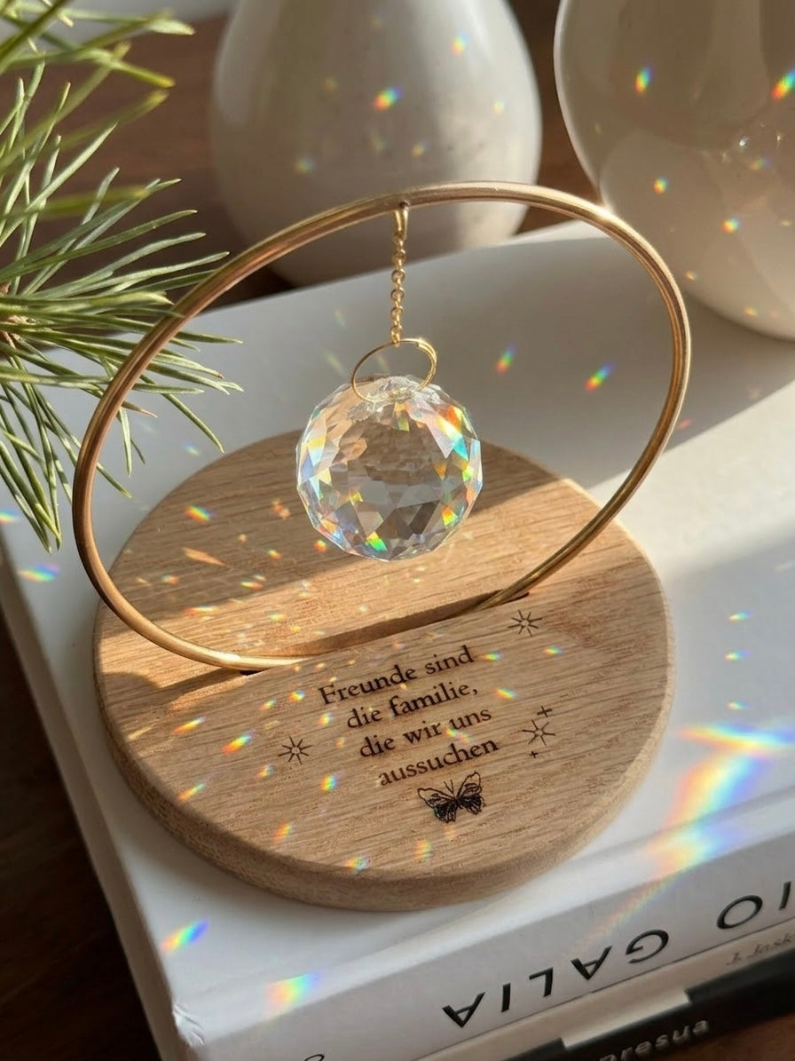 The Circle - A minimalist Sonnenfänger (suncatcher) featuring a high-quality faceted Kristall-Prisma suspended by a delicate gold stainless steel chain within a matching gold ring. The modern tabletop design rests on a solid, dark redwood base (Echtholz-Sockel) and is shown creating vibrant rainbow light reflections (Regenbogen-Effekt) across the background. This elegant Fensterdeko is perfect as a mindful Geschenkidee for home or office.
