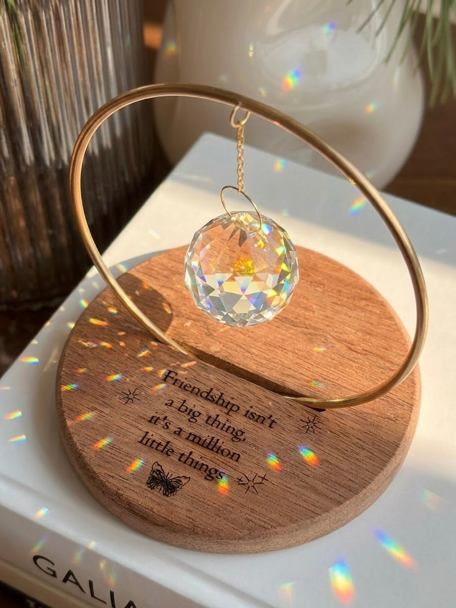 The Circle - A minimalist Sonnenfänger (suncatcher) featuring a high-quality faceted Kristall-Prisma suspended by a delicate gold stainless steel chain within a matching gold ring. The modern tabletop design rests on a solid, dark redwood base (Echtholz-Sockel) and is shown creating vibrant rainbow light reflections (Regenbogen-Effekt) across the background. This elegant Fensterdeko is perfect as a mindful Geschenkidee for home or office.