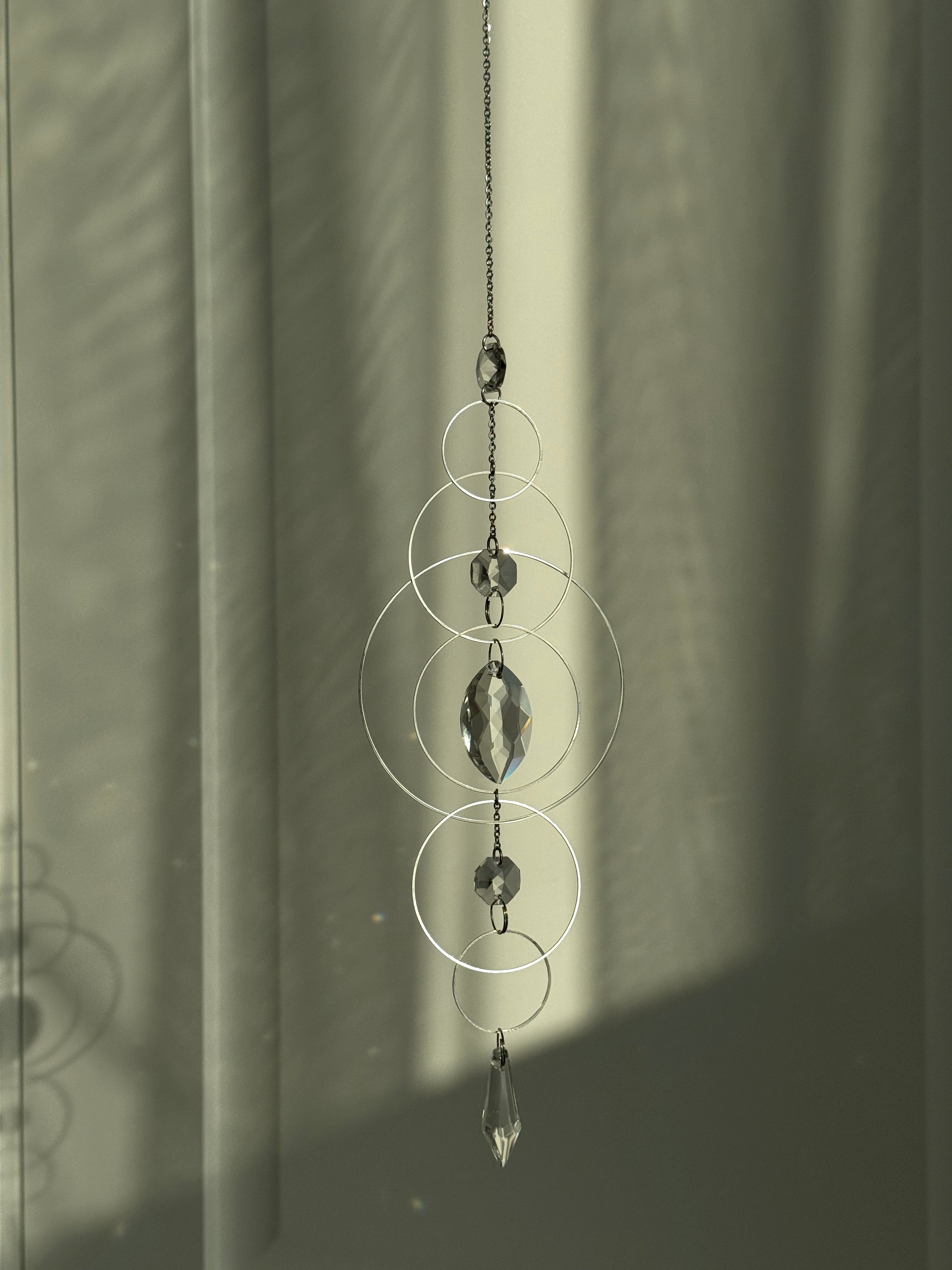 60cm silver geometric suncatcher featuring interlocking concentric hoops and clear marquise-cut crystal prisms, hanging in a window and casting rainbows on a wall.