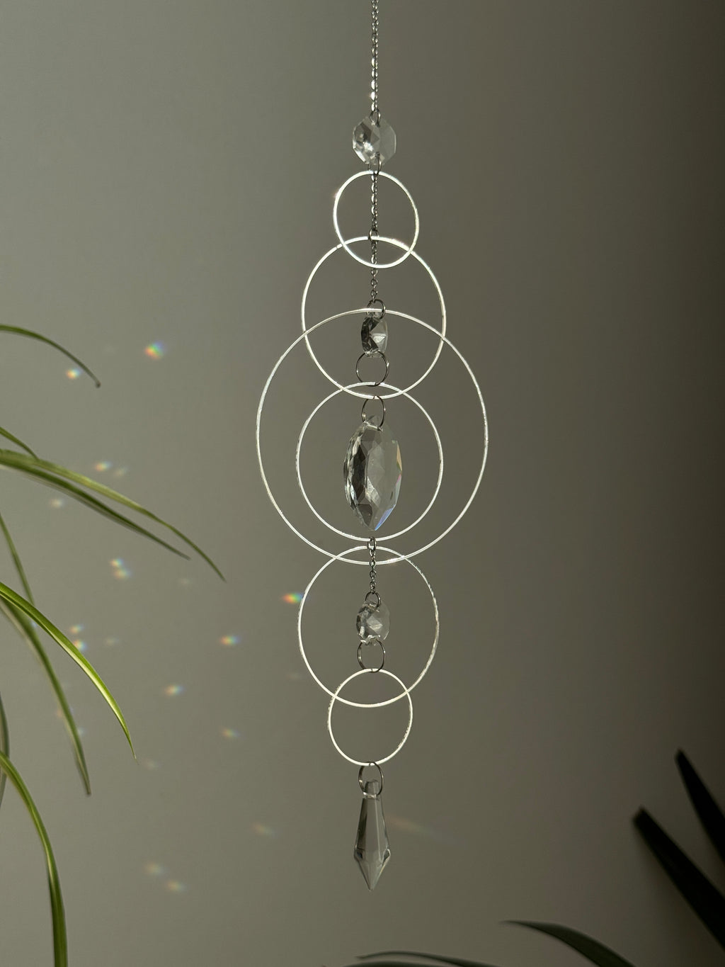 60cm silver geometric suncatcher featuring interlocking concentric hoops and clear marquise-cut crystal prisms, hanging in a window and casting rainbows on a wall.