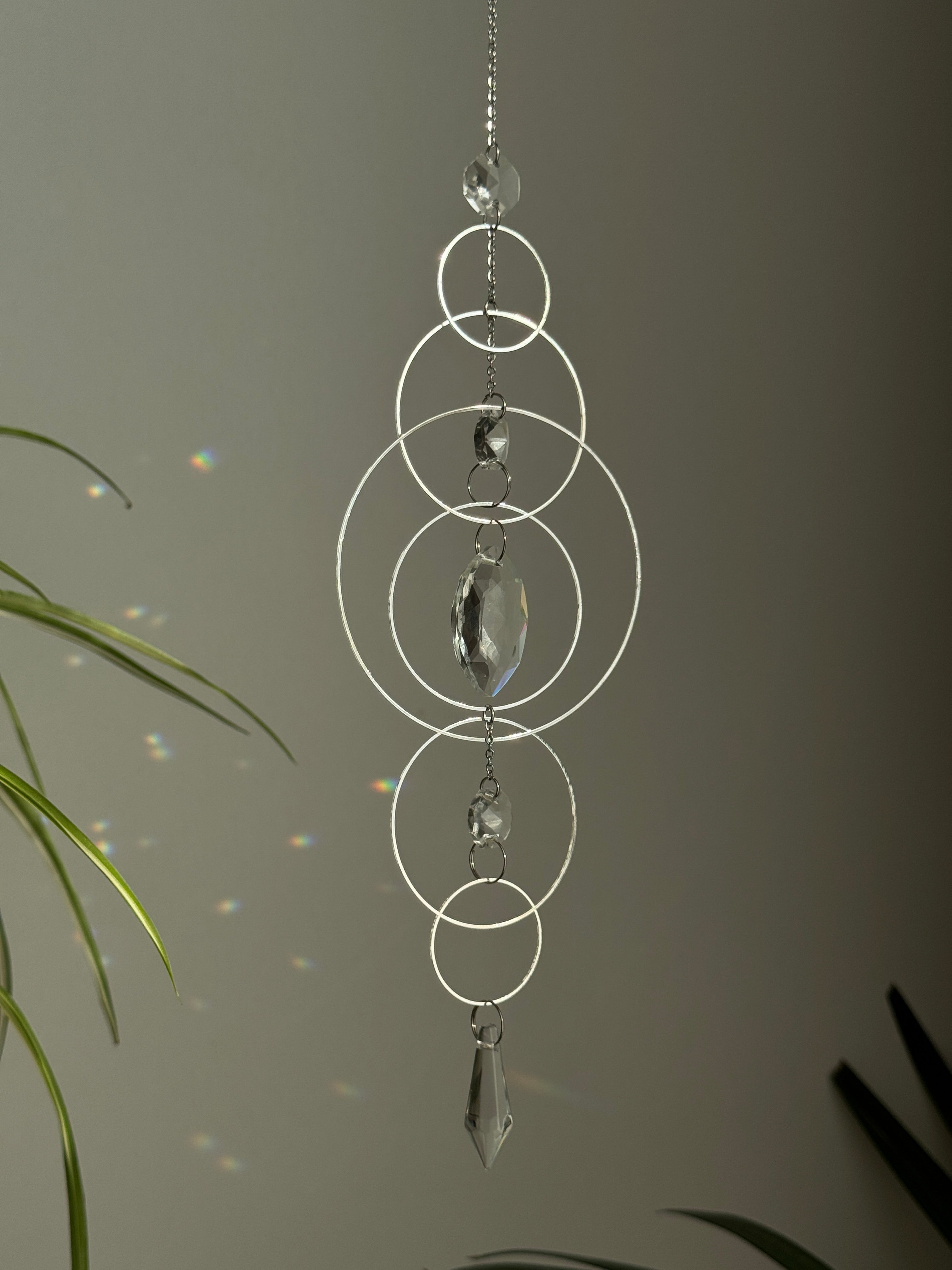 60cm silver geometric suncatcher featuring interlocking concentric hoops and clear marquise-cut crystal prisms, hanging in a window and casting rainbows on a wall.