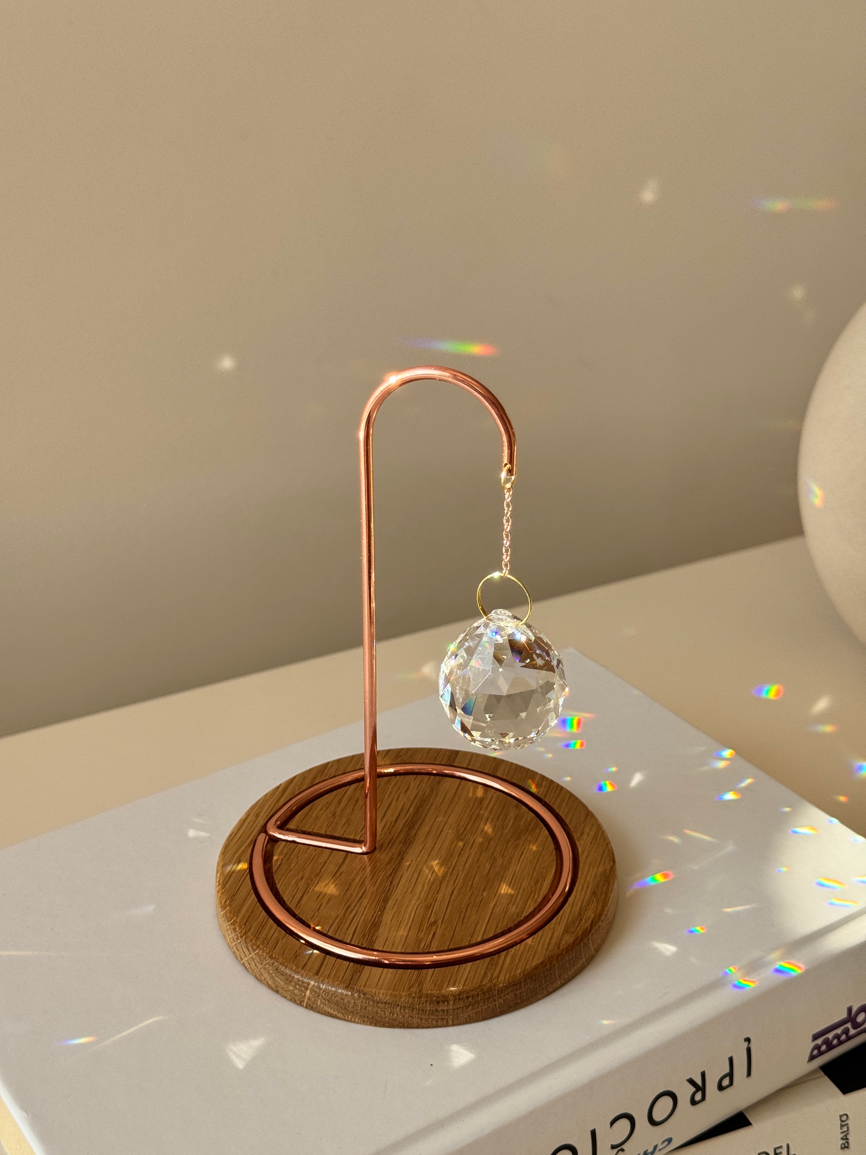 A close-up photograph of a clear, multi-faceted crystal suncatcher prism hanging from an ornate rose gold metal hook on a dark wooden base. The suncatcher casts multiple rainbow-colored refractions of light across the white tabletop, a stack of white books on the left, and a textured off-white ceramic vase in the background. Dried branches are visible emerging from the vase against a light gray wall.
